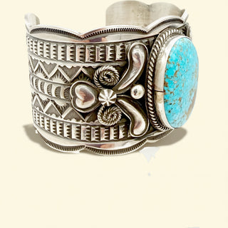 Silver cuff bracelet with intricate designs and a turquoise stone on a beige background