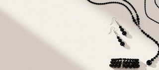 Black beaded jewelry set including a necklace, drop earrings, and a bracelet, arranged on a light beige surface with soft diagonal shadows.