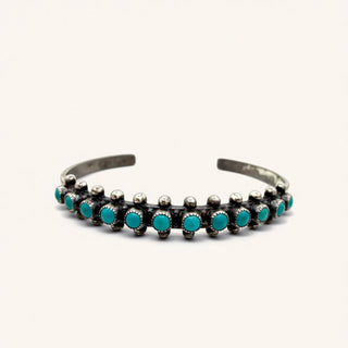 A sterling silver cuff bracelet featuring a row of turquoise stones in oval bezel settings, separated by small silver beads, on a light cream background.