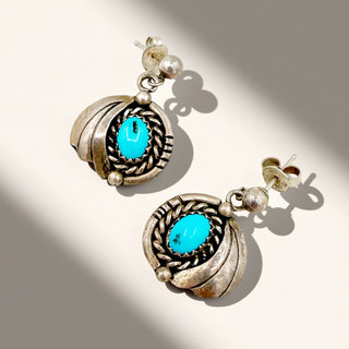 A pair of silver-toned drop earrings with oval turquoise cabochons and small pearl-like beads, photographed on a light beige background with strong diagonal shadows.