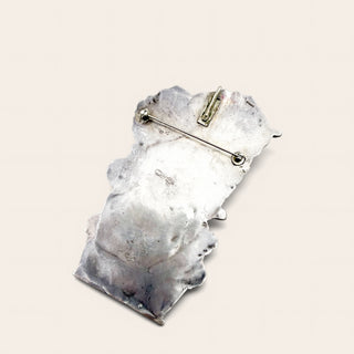 A textured silver pin or brooch with an irregular organic shape and a safety pin clasp, shown on a light beige background.