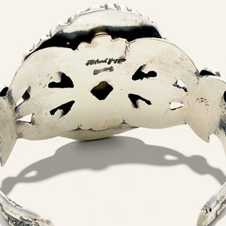 A sterling silver cuff bracelet with a textured, organic shape and cutout details, featuring an engraving on the inner band, shown against a light beige background.