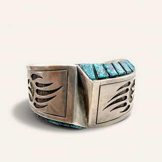 A wide sterling silver cuff bracelet featuring turquoise stone inlay and engraved geometric and wing-like cutout details, shown on a light beige background.