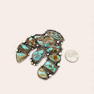 A Southwestern-style silver pendant featuring multiple turquoise and brown stone clusters, displayed next to a US quarter coin for scale on a light beige background.