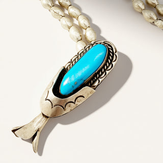 A close-up of a silver-toned pendant necklace with a bright turquoise blue oval stone, shown against a light cream background with part of the white beaded chain visible at the top.