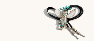A silver-toned Southwestern-style BOLO TIE with turquoise and white stone inlay, featuring a central feathered motif and braided black cords, photographed on a clean off-white background.
