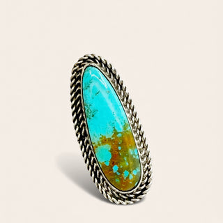 A silver-toned rope-textured ring featuring an oval turquoise stone with natural brown matrix and blue-green coloration, set against a light beige background.
