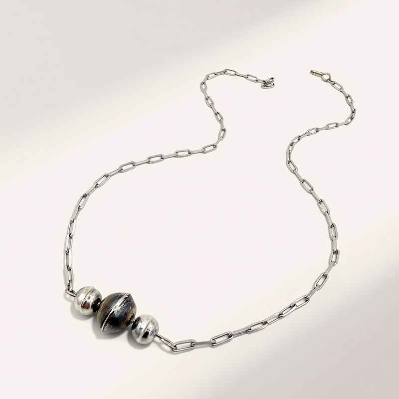 A silver chain necklace featuring three polished metallic beads, including one larger central bead with a dark reflective center, displayed on a clean white background.