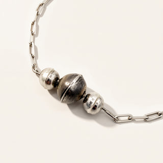 A close-up of a silver chain necklace featuring three metallic spherical beads, with one larger textured bead in the center and two smaller polished beads on either side, set against a plain off-white background.
