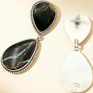 Teardrop-shaped earrings with black and green stone and silver frame on a beige background