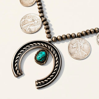 A silver-toned beaded necklace with a large open crescent pendant featuring a turquoise stone, displayed on a light beige background with several silver coins arranged nearby.