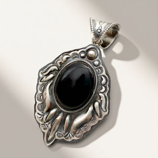A silver pendant with an ornate handcrafted design featuring a large oval black cabochon stone in the center, set against a clean, light beige background.