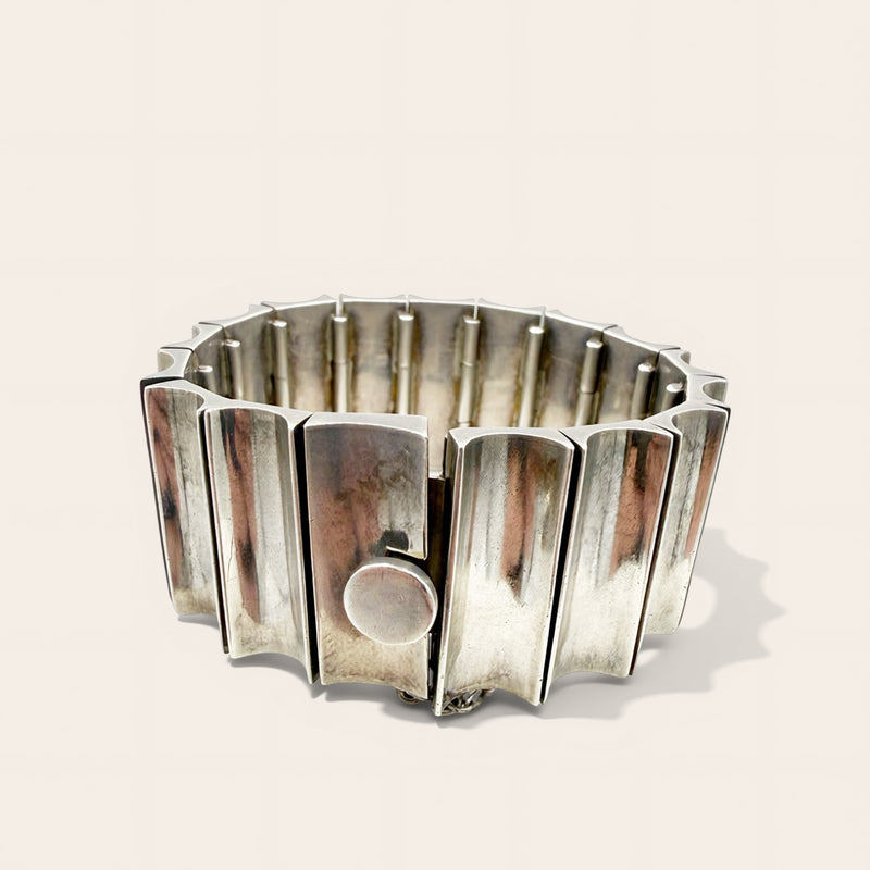 A wide, chunky silver metal cuff bracelet with a segmented, flared design and a circular clasp, photographed on a light beige background.
