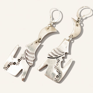 A pair of sterling silver dangle earrings shaped like stylized cat heads, featuring textured details and circular ear loops, displayed on a light cream background.