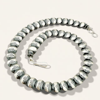 A sterling silver beaded necklace with flat, round disc beads featuring engraved floral or leaf-like patterns, arranged in a spiral on a light beige background.