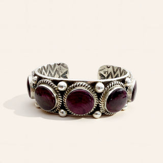A silver cuff bracelet featuring three large oval dark purple gemstones and smaller side stones, set in a textured, handcrafted sterling silver design on a light beige background.
