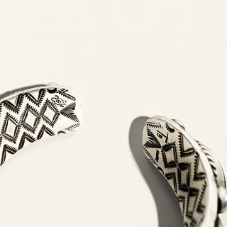 Close-up of a silver cuff bracelet with a black geometric pattern, shown on a warm off-white background with a soft shadow.