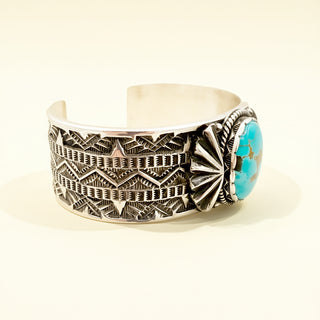 Silver cuff bracelet with intricate patterns and a turquoise stone on a light background