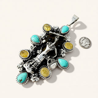 A silver-toned Southwestern-style pendant featuring turquoise and yellow gemstone accents, displayed on a light beige background with a coin for scale.