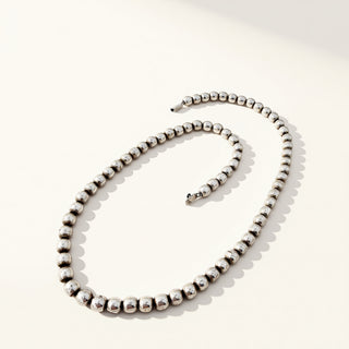 A silver beaded necklace with a textured, polished finish, arranged in a loose spiral on a clean, off-white background.