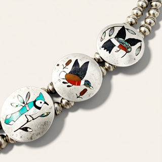 A close-up of a silver beaded necklace featuring three round inlay discs decorated with colorful bird designs in turquoise, black, red, and white, set against a light beige background.