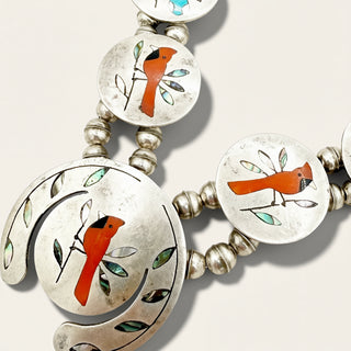 Silver-toned beaded necklace with oval and crescent-shaped inlay discs featuring red cardinal birds and green-and-white leaf motifs on a light beige background.