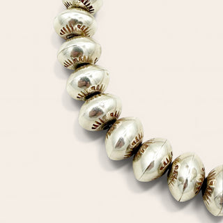 A close-up of a metallic beaded strand, likely sterling silver jewelry components, featuring polished oval beads with engraved floral details, set against a solid light beige background.