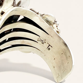 Close-up of a silver-toned curved decorative object with layered curved bands and engraved markings, photographed on a light neutral background.