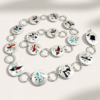A silver-toned chain bracelet arranged in a spiral on a light beige background with soft shadows. Each oval link features a colorful abstract design in black, red, teal, and white.