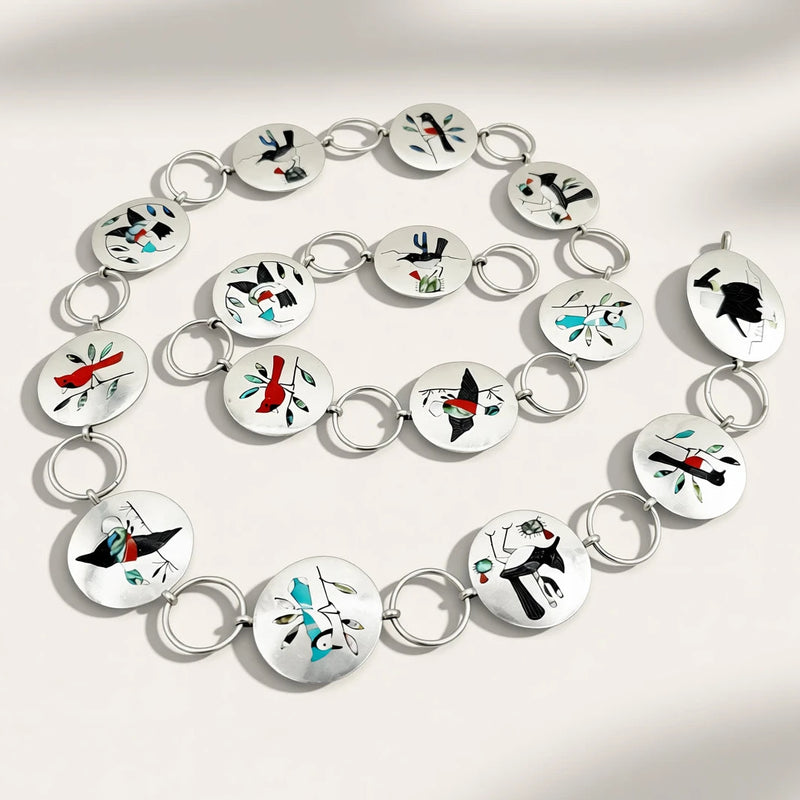 A silver-toned chain bracelet arranged in a spiral on a light beige background with soft shadows. Each oval link features a colorful abstract design in black, red, teal, and white.