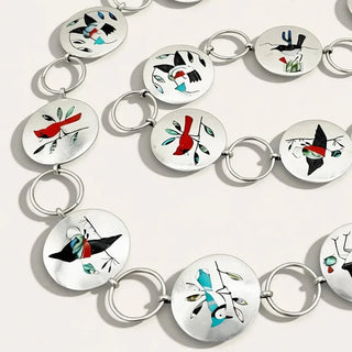 A silver-toned circular link chain necklace featuring multiple round white inlaid discs decorated with abstract black, red, and turquoise geometric and bird-like motifs, arranged on a light neutral background.