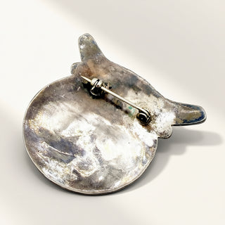 A silver-toned metal brooch shaped like a stylized bird with a circular base and a pin clasp, photographed on a plain light background.