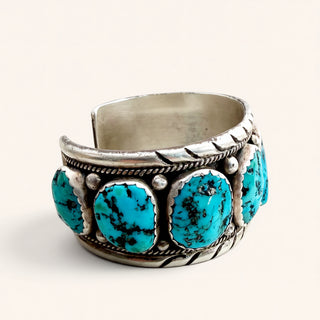 A sterling silver cuff bracelet featuring five oval turquoise stones, photographed on a clean, light beige background.