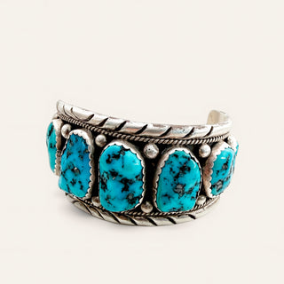 A sterling silver cuff bracelet featuring five oval turquoise stones, set against a plain off-white background.