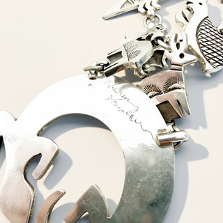 A close-up of a silver-toned jewelry piece featuring a large crescent-shaped pendant with engraved script, accompanied by small charm elements on a light neutral background.