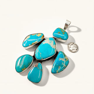 A sterling silver pendant featuring seven polished turquoise stones of various shapes, displayed on a clean off-white background with a small coin for scale.