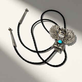 A black braided rope bolo tie with a detailed silver-toned metal central charm featuring a turquoise blue stone, displayed on a light gray background with artistic diagonal shadows.