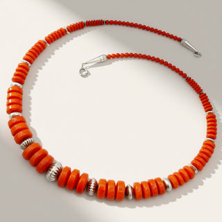 A beaded necklace with vibrant orange-red cylindrical beads and silver-toned spacer beads, arranged in a circle on a light gray background.