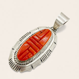 A sterling silver pendant featuring a carved red coral cabochon with geometric patterns, set in a scalloped silver bezel with a bail at the top, shown on a light beige background.