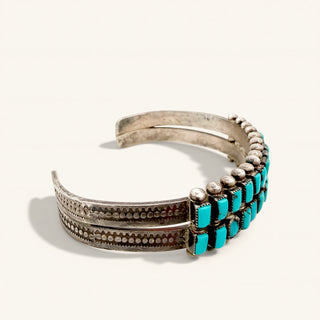 A silver-toned Southwestern-style cuff bracelet decorated with rows of vibrant turquoise stones and small metallic beads, set against a plain off-white background.