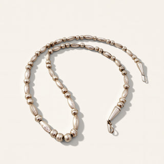 A silver-toned beaded chain necklace with elongated oval beads and smaller round beads, displayed on a light cream background.