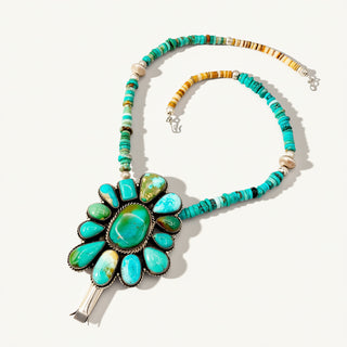 A Southwestern-style turquoise necklace featuring a large floral cluster pendant with turquoise and green agate stones in silver bezel setting, connected to a beaded chain with turquoise, brown, and cream beads on a light beige background.