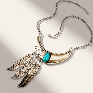 A silver-toned statement necklace featuring a crescent moon and feather pendant design with a turquoise-colored oval stone, displayed on a light beige background with soft shadows.