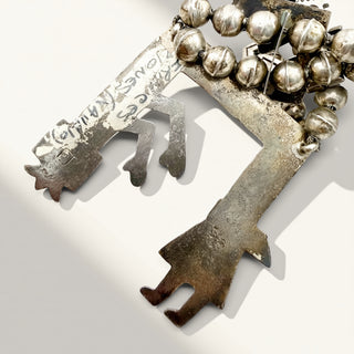 A rustic silver-toned metal key-shaped pendant or ornament with engraved markings, a textured patina, and a cluster of small round bells along the top, photographed on a light gray background.