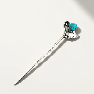A silver-toned hair pin or hair stick with a turquoise blue stone and a leaf-shaped detail, photographed on a clean off-white background with a soft shadow.