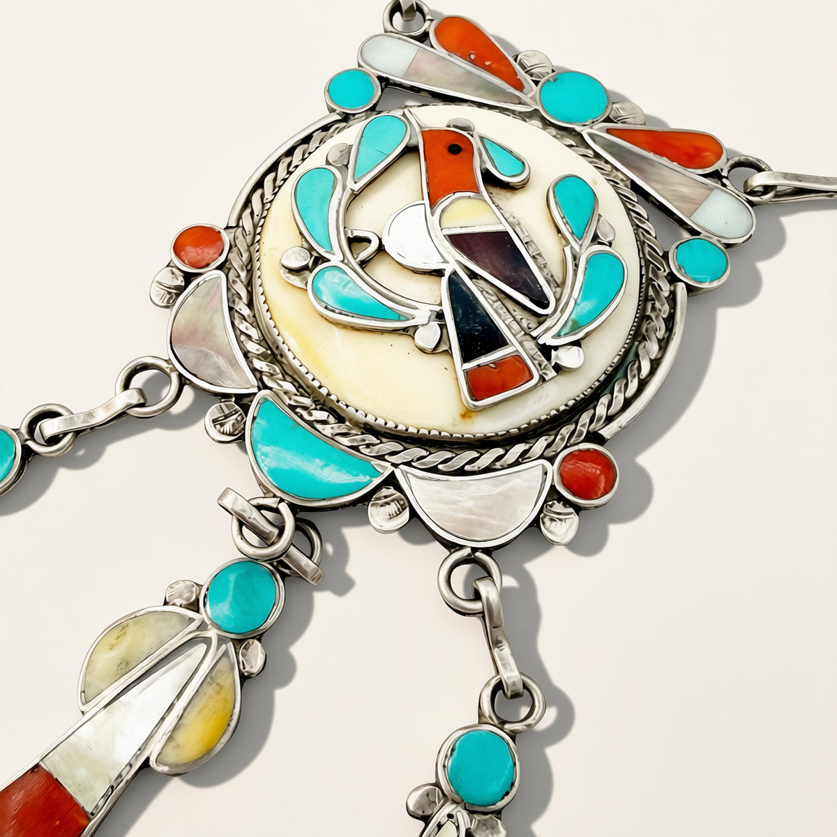 A close-up of a silver-toned Southwestern-style jewelry piece featuring turquoise, coral, white shell, and dark stone inlay details on a light background.