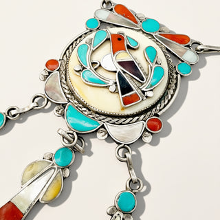 A close-up of a silver-toned Southwestern-style jewelry piece featuring turquoise, coral, white shell, and dark stone inlay details on a light background.