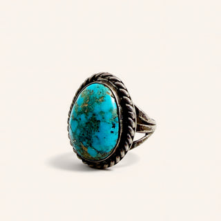 A silver ring featuring a large oval turquoise cabochon stone with natural matrix patterns, set in a braided rope-style bezel on a light cream background.