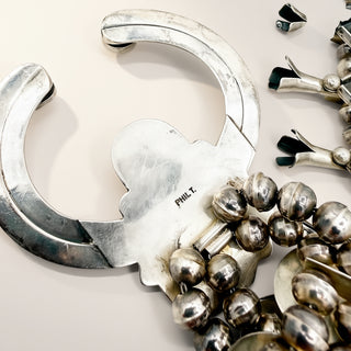 A close-up of a silver-toned jewelry piece featuring a large open C-shaped curved section, a central decorative emblem, and multiple dangling round silver beads. The back of the piece is stamped with the brand name 'PHIL.