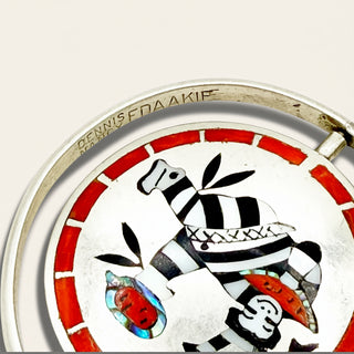 A close-up of a circular silver pendant featuring hand-inlaid Native American style designs, including black and white striped figures, red accents, and a blue iridescent shell piece. The inner rim is engraved with the name 'DENNIS FDAAKIE'.